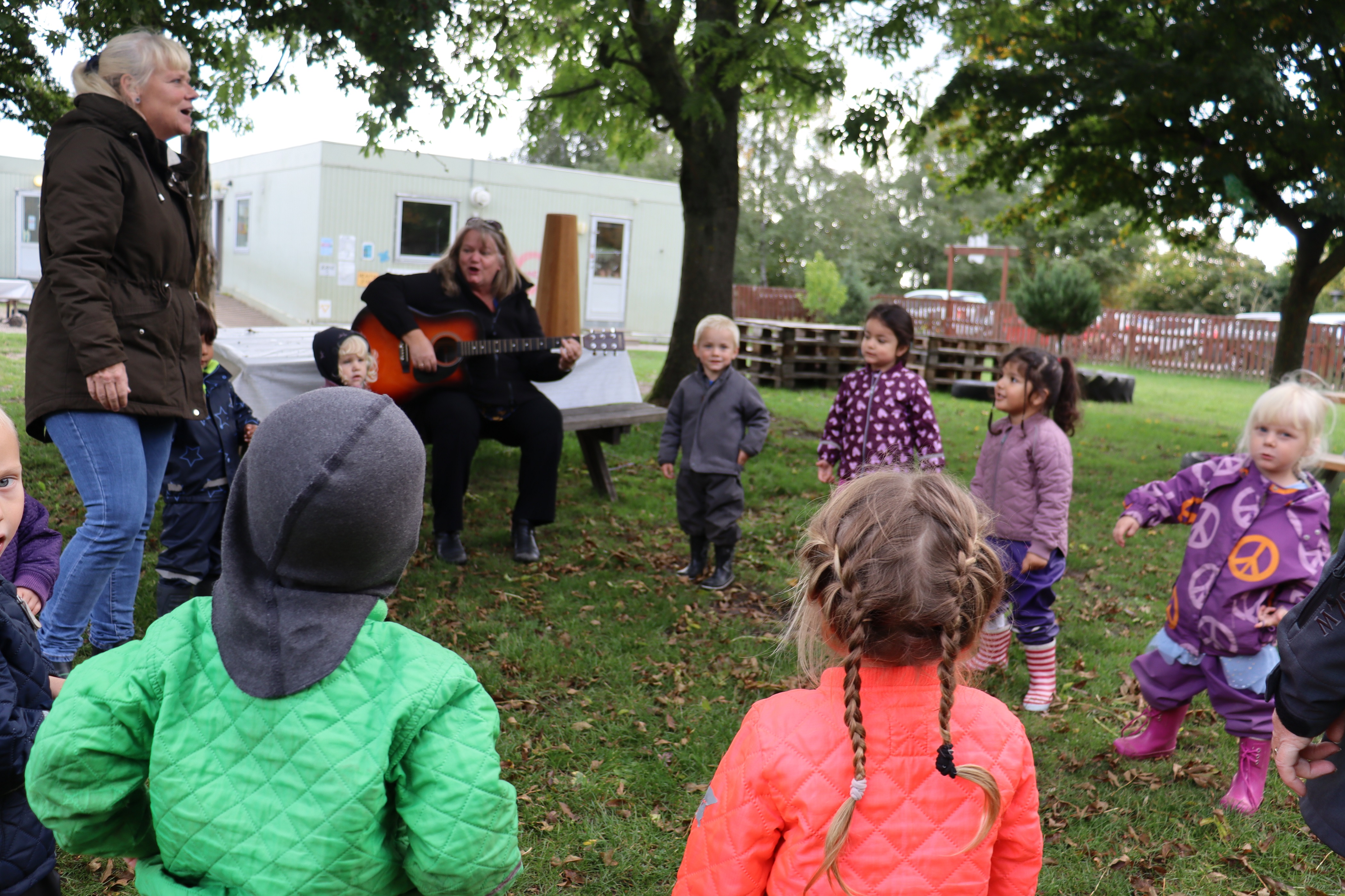 Pædagog spiller guitar udenfor, hvor børn er samlet i en rundkreds omkring